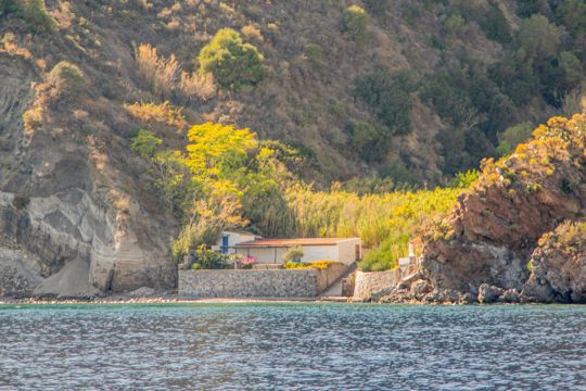 A small waterside building beneath a green hillside and cliffs